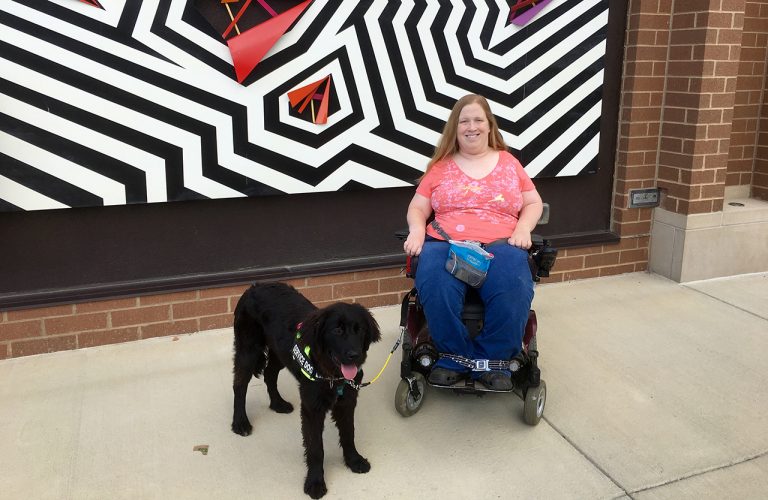 Karin Willison with my service dog sitting in my wheelchair outside in front of a zigzag mural.